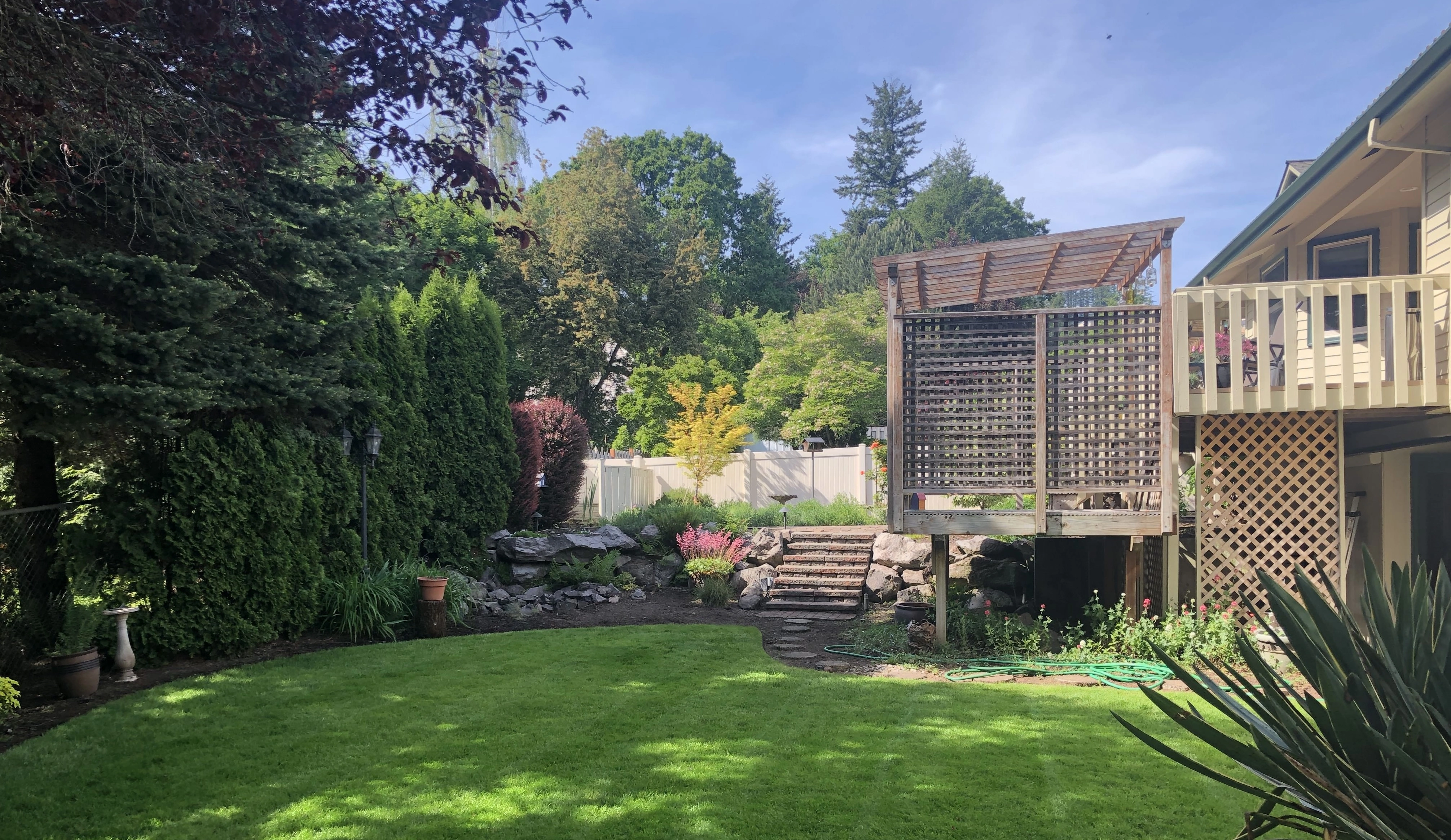 A backyard with a green lawn, garden, trees, and a wooden deck with lattice railing in Gresham, OR.